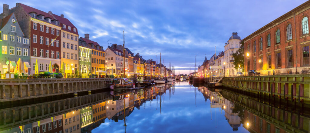 Panoramic view of Nyhavn in Copenhagen, Denmark, featuring colorful facades of old houses, traditional ships docked along the canal, and the historic charm of the old town.